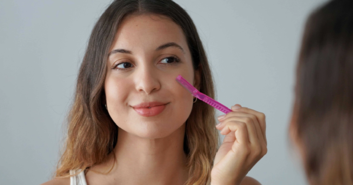 A young woman in Humble, TX, smiling while using a pink dermaplaning tool on her cheek in front of a mirror.